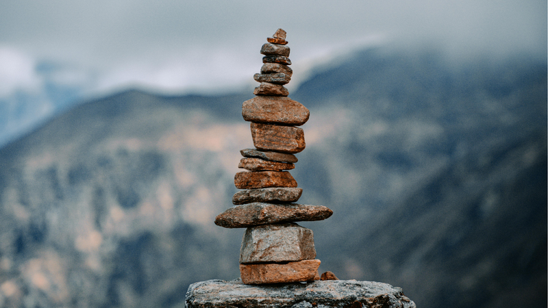 A balanced stack of natural stones, or cairn, standing on a rock with a blurred mountain range in the background under a cloudy sky.